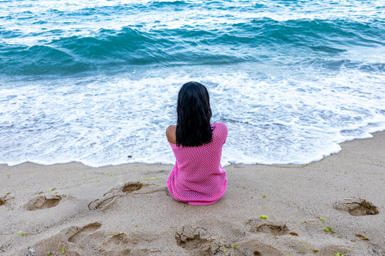 Young girl in pink dress sitting alone facing the ocean, waves gently rolling in on sandy shore under a cloudy sky.