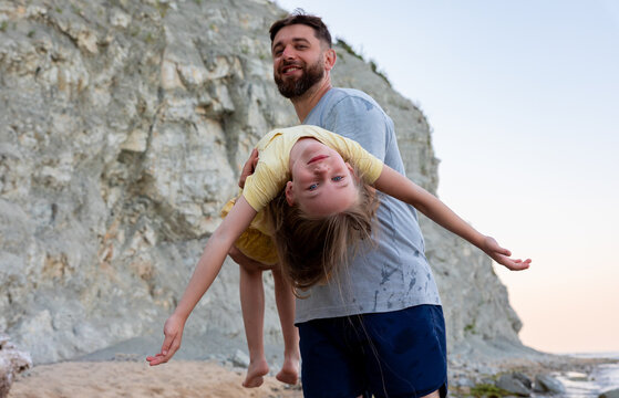Smiling father holding his daughter upside down on a scenic rocky beach,  enjoying playful family moments against a natural cliff backdrop.