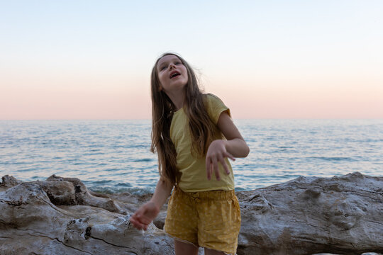 Young girl laughing by the sea at sunset, standing on driftwood with calm waves and soft pastel sky in the background.