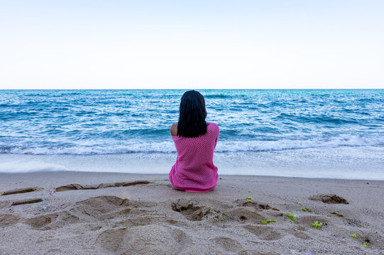 Rear view of a woman sitting alone on sandy beach facing the calm ocean under clear sky, wrapped in a pink shawl, evoking peace and solitude.