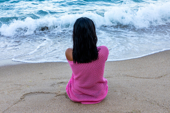 Rear view of woman in pink top sitting on sandy beach facing ocean waves, evoking relaxation and summer vacation concepts.