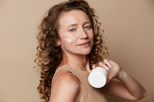 Authentic beauty of a middle-aged woman with natural skin without retouching. She has curly hair and holds a cream jar, applying lotion on her cheek. Studio shot on a beige background.