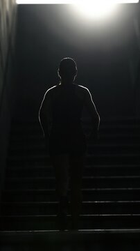 Silhouette of a woman in athletic wear walking up dark stairs illuminated by bright overhead light