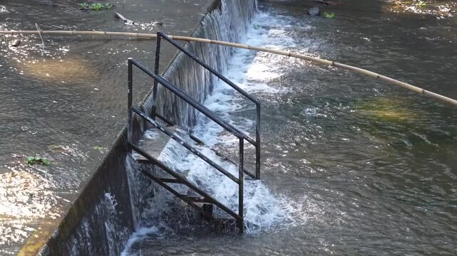 A black metal staircase submerged in water at a small river dam with flowing rapids.