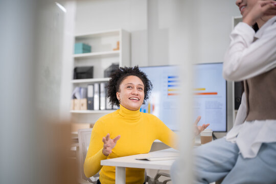 Cheerful professional businesswoman in a yellow sweater talking and gesturing during a team meeting in a modern corporate office setting.