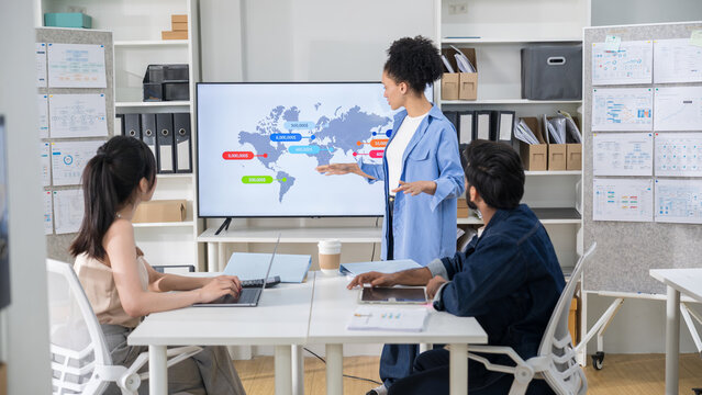 Female executive leading a corporate meeting, presenting global business data and a world map on a digital screen to her seated team members.