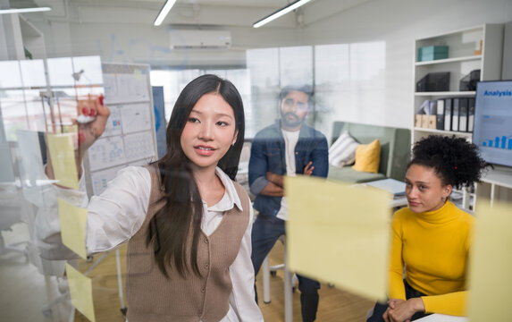 Professional businesswoman writing on a glass board with colorful sticky notes during a collaborative project planning session with her team.