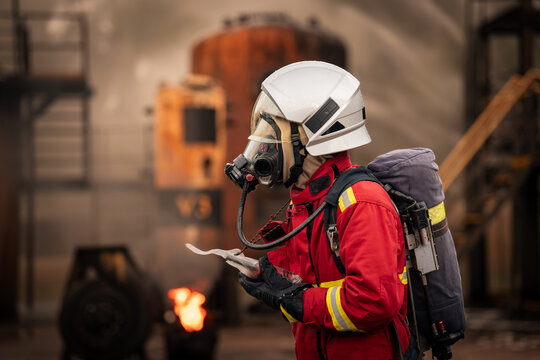 Firefighter Using a Walkie Talkie beside Industrial Flames while Wearing a Full Face Mask, White Helmet, and Red Protective Uniform