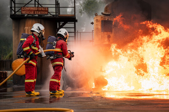 Firefighter Team Operating a Fire Hose beside Large Industrial Flames while Wearing Full Protective Uniforms and Air Tanks