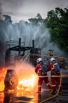 Firefighter Team Advancing toward Industrial Flames with a Fire Hose and Air Tanks across Wet Pavement under Water Spray