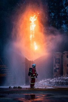 Firefighter Standing near a Burning Industrial Structure amid Water Spray while Wearing a White Helmet, Red Uniform, and Air Tank