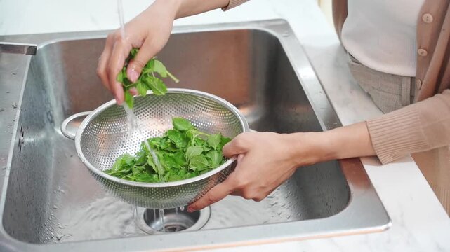 Woman washing fresh green leafy vegetables in metal colander under kitchen sink faucet water