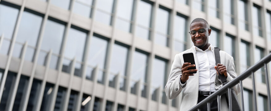 Black businessman, using smartphone, texting, works as accountant, outdoor near office building. African american business man smiling, committed to career growth, innovating. Copy space. Web banner.