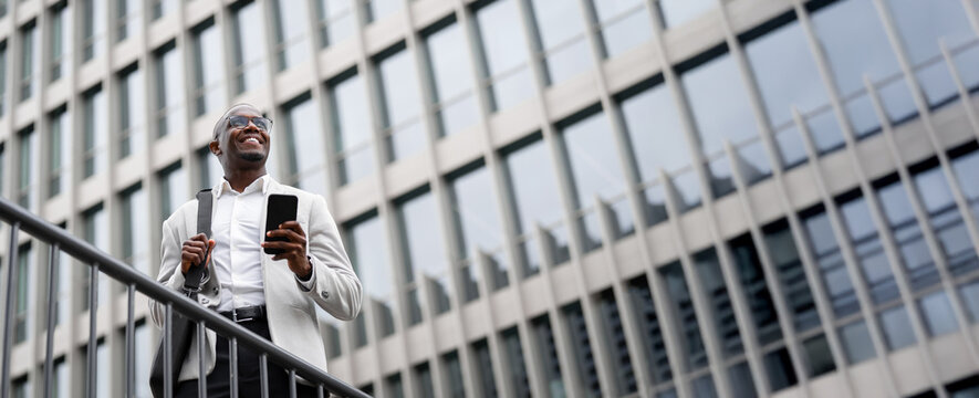 Financial advisor african american businessman outdoor near office building, holding smartphone, looking aside. Black business man smiling, managing work remotely, innovating. Copy space. Web banner.