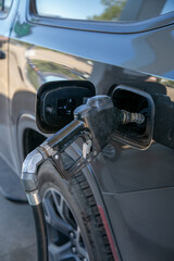 Close-up of a fuel nozzle inserted into the filler neck of a dark grey vehicle during refuelling. The open fuel door reveals the fuel cap and filler area while petrol or diesel is dispensed © Val