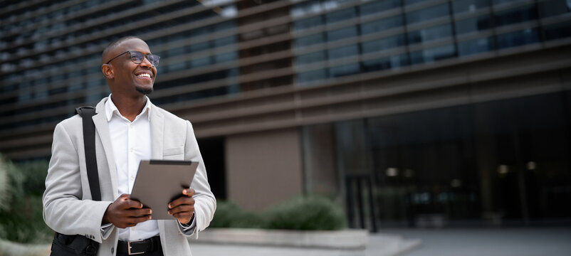 Business consultant black businessman outdoor near office building, holding tablet, looking aside. African american business man smiling, staying connected with technology. Copy space. Web banner.