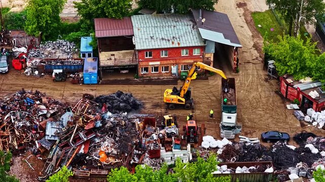 Aerial view of an industrial scrap metal yard with an excavator loading a truck, surrounded by piles of metal and industrial buildings in Latvia.