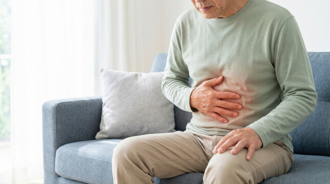 Older man stomach pain digestive problem abdominal discomfort senior health home interior sitting casual clothing living room healthcare concept pain illness disease symptom ache Asian male sofa