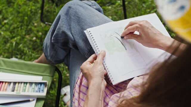 Caucasian woman drawing with pastel crayons in sketchbook outdoors, over shoulder view. Footage ideal for promoting art therapy, creative hobbies, and outdoor recreation campaigns