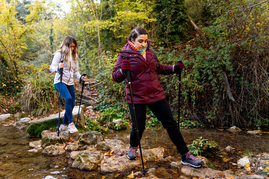 Two women enjoying nordic walking in autumn nature, crossing a stream with poles
