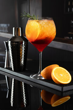 Citrus cocktail with rosemary and ice in glass, shaker, jigger and orange halves on black bar mat. Aperol on dark background. Sprig of rosemary
