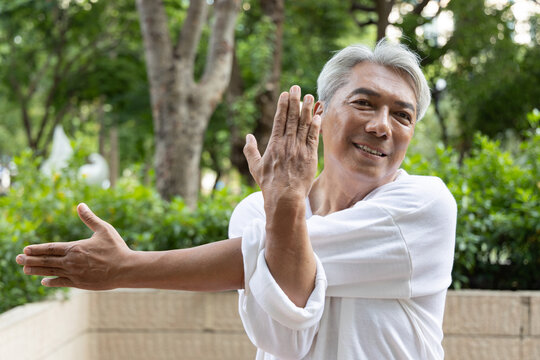 Happy smiling asian senior man stretching his shoulder and back muscles during an outdoor workout in an urban park, healthy aging concept