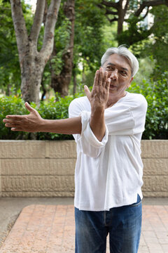 Awkward asian senior man trying to stretch his shoulder and back muscles during an outdoor workout in an urban park, the beginning of healthy aging concept