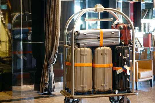 Luggage cart holds multiple suitcases in elegant hotel lobby. Shiny metallic cases gleam under warm ambient lighting. Curled curtains frame the reflective glass doors
