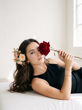 A Woman's Delicate Touch: A young woman with a floral adornment in her dark, wavy hair reclines gracefully, holding a deep red rose delicately against her eye.