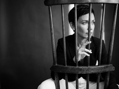 Contemplation Behind Bars: A woman with a striking short haircut sits inside a wooden chair, her gaze directed away as if lost in thought, creating a powerful visual of introspection and confinement.