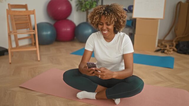 Woman holding smartphone seated crosslegged on a yoga mat in an exercise studio, smiling and checking phone, wearing t shirt and leggings; serenity wellbeing.
