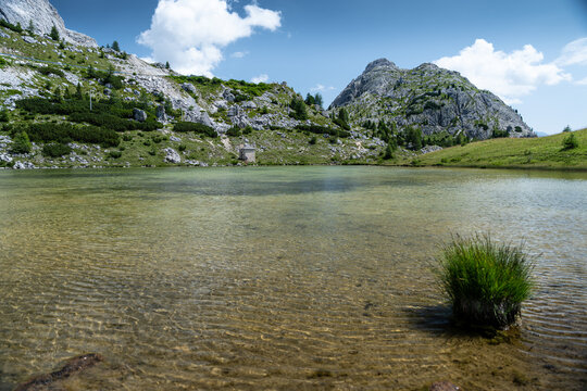 View of the clear waters of Lago di Valparola at Passo Valparola, surrounded by rocky mountain peaks, green alpine slopes, and a bright blue sky in Cortina d'Ampezzo, Veneto, Italy.