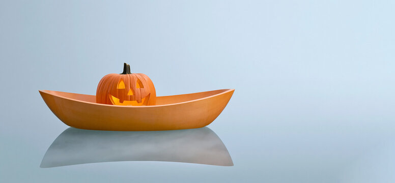 Pumpkin regatta. Carved Halloween pumpkin with a smiling face placed inside an orange bowl on a reflective surface against a light blue background