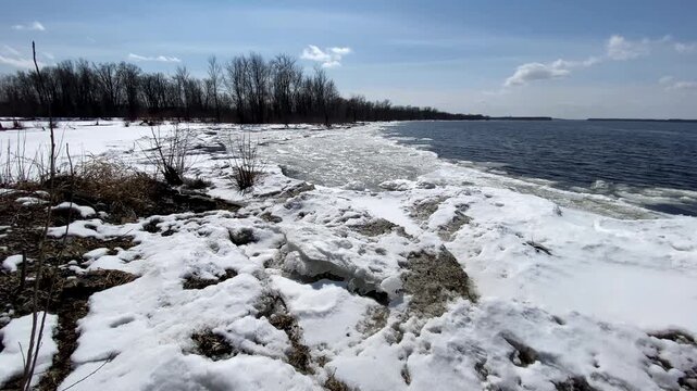 Melting ice floes and snow piled along a shoreline under a clear blue sky, bordering open dark blue river water during early spring thaw.