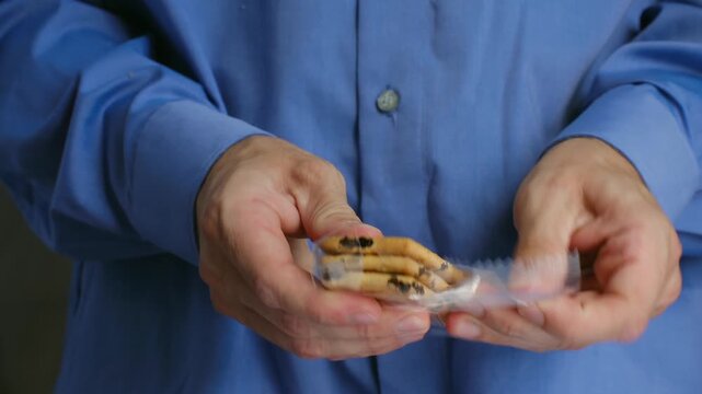 Close up man hands wearing blue shirt opening transparent plastic package raisin crackers, getting ready for quick and convenient snack during work break at office. Businessman opening raisin crackers