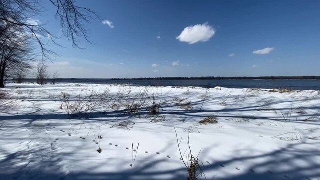 Melting ice floes and snow piled along a shoreline under a clear blue sky, bordering open dark blue river water during early spring thaw.