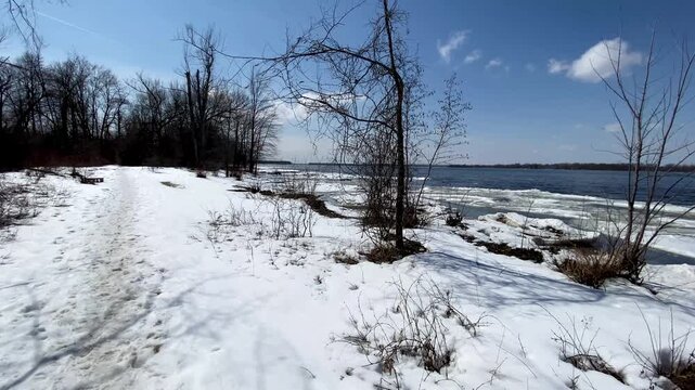 Snow-covered riverbank with bare trees and melting ice floes floating along the open blue water under a clear sunny sky.