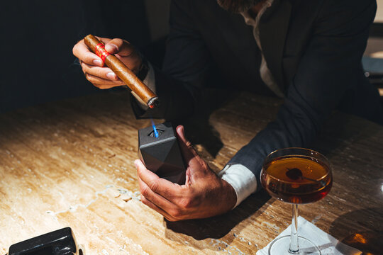 Businessman Lighting Cigar With Modern Lighter at Bar Table Beside Cocktail Glass and Ashtray