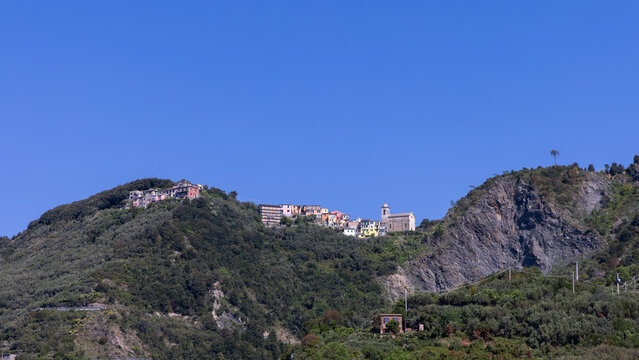 Corniglia by Ligurian waters in Italy: Small clustered houses and a church line a green ridge beneath a large cloudless sky.