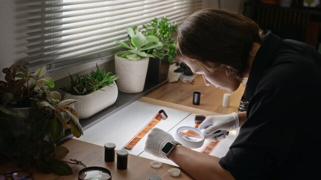 Young Asian photographer wearing gloves inspecting film negatives with magnifying glass on light box. Clip is ideal for analog photography workshops and creative studio marketing