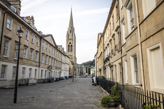 View of a historic street leading towards a church spire, featuring traditional stone architecture, a cobblestone path, and a street lamp in Bath, England, United Kingdom.