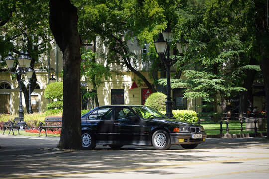 Three-quarters profile view of a youngtimer car black BMW 3 Series (E36) sedan parked in a green shaded park area in center of Subotica, Serbia, on June 7, 2026