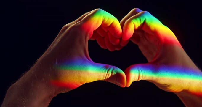 Close-up studio video of two hands forming a heart shape illuminated by rainbow-colored sunlight against a black background. Symbolic LGBTQ+ Pride concept representing love, equality, diversity