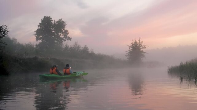 Two adventurous people wearing life jackets paddle kayak on calm, misty river during foggy sunrise. Surrounded by serene and tranquil natural landscape. Travelers are filming fabulous sunrise on phone