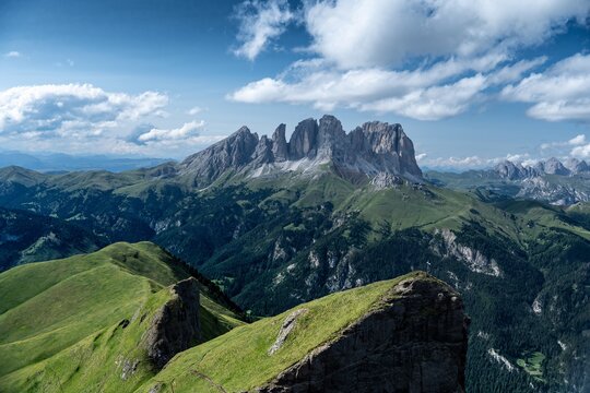 View of the Crepa Neigra mountain range with lush green alpine meadows and rocky peaks under a blue sky in Canazei, Trentino-Alto Adige, Italy.