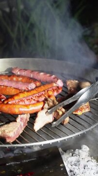 A close-up shot of a home barbecue with a realistic nighttime look, featuring chorizo, sausages, and steaks. A steel tong is flipping a piece of meat.