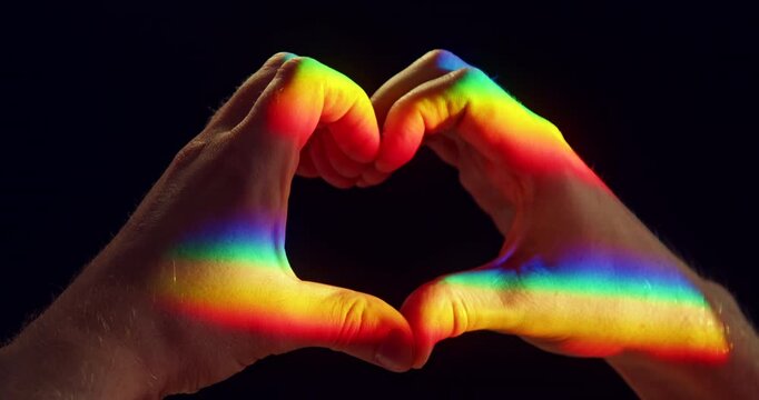 Close-up studio video of two hands forming a heart shape illuminated by rainbow-colored sunlight against a black background. Symbolic LGBTQ+ Pride concept representing love, equality, diversity