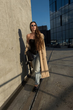 Confident young woman walking near a concrete wall on a sunny city street. Stylish fashionable female model in beige trench coat, sunglasses, and jeans outdoors. Modern urban lifestyle photo.