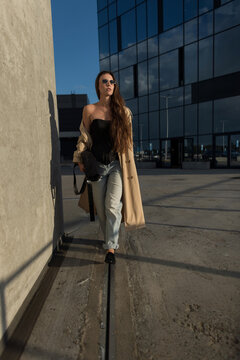 Stylish young woman walking confidently near modern glass building. Fashionable model wearing beige trench coat, sunglasses, and jeans outdoors. Trendy urban street style lifestyle photo.