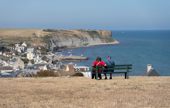 Arromanches-les-Bains, France - 08 11 2026: The Garden of Remembrance. An elderly couple sitting on a bench, looking the sea, the beach, the city, cliffs and coastal path
